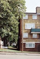 A residential scene with a brick apartment building featuring several windows, a green balcony, and a road sign that reads 'Dacre Park'. A tree with lush green foliage partially obscures the building. A street runs along the foreground, and a car is visible parked in the background. The atmosphere is quiet and suburban.