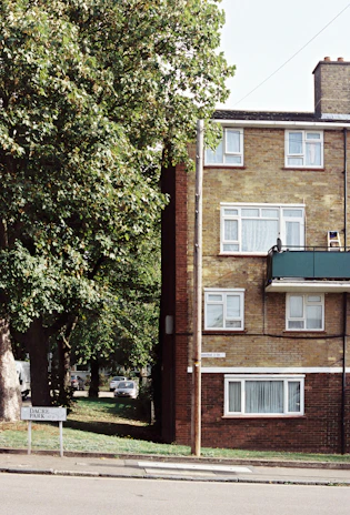 A residential scene with a brick apartment building featuring several windows, a green balcony, and a road sign that reads 'Dacre Park'. A tree with lush green foliage partially obscures the building. A street runs along the foreground, and a car is visible parked in the background. The atmosphere is quiet and suburban.