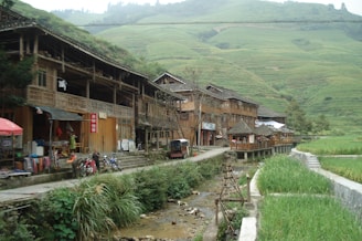 A rural village scene featuring traditional wooden houses built along a small stream. The buildings have open fronts displaying various goods, indicating market activity. Several motorcycles are parked along the pathway, which runs parallel to the stream. In the background, terraced hillsides stretch up the hills covered with lush green vegetation.