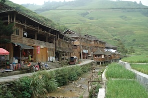 A rural village scene featuring traditional wooden houses built along a small stream. The buildings have open fronts displaying various goods, indicating market activity. Several motorcycles are parked along the pathway, which runs parallel to the stream. In the background, terraced hillsides stretch up the hills covered with lush green vegetation.