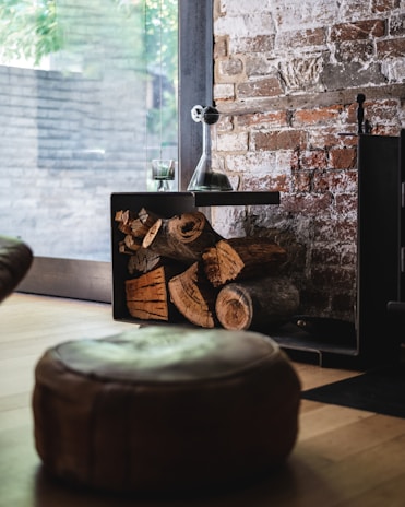 A cozy indoor setting featuring a rustic brick wall and a window with a view of green foliage outside. There is a stack of firewood neatly arranged within a black metal holder beside a fireplace. A clear glass decanter and a matching glass sit on top of the holder, reflecting a relaxed ambiance. A leather pouf is positioned in the foreground on a light wooden floor.
