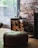 A cozy indoor setting featuring a rustic brick wall and a window with a view of green foliage outside. There is a stack of firewood neatly arranged within a black metal holder beside a fireplace. A clear glass decanter and a matching glass sit on top of the holder, reflecting a relaxed ambiance. A leather pouf is positioned in the foreground on a light wooden floor.
