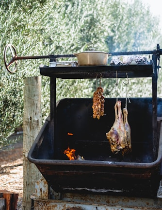 A rustic outdoor cooking setup with a rotisserie system is pictured. Meat is being roasted, with a skewer of skewered pieces and a larger slab of meat hanging inside a metal container. There is a pot on a metal grate above the meat. A small flame is visible below, and the background shows dense, leafy vegetation.