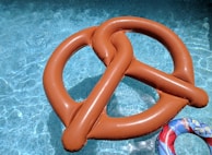 A large, inflatable float in the shape of a pretzel is situated in a swimming pool with clear blue water. The sun casts reflections on the pool's surface, enhancing the vibrant appearance of the scene. A smaller, colorful pool ring with images on it is partially visible in the bottom right corner.