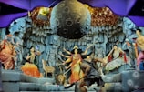 A vibrant depiction of a mythological scene featuring various colorful statues and intricate details. Central figures include deities with multiple arms, a figure resembling an elephant, and other humanoid characters. The backdrop resembles a rocky cave with a large celestial object suspended above. Decorative elements and lighting add to the drama and complexity of the setting.