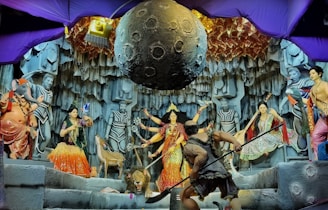 A vibrant depiction of a mythological scene featuring various colorful statues and intricate details. Central figures include deities with multiple arms, a figure resembling an elephant, and other humanoid characters. The backdrop resembles a rocky cave with a large celestial object suspended above. Decorative elements and lighting add to the drama and complexity of the setting.
