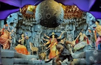 A vibrant depiction of a mythological scene featuring various colorful statues and intricate details. Central figures include deities with multiple arms, a figure resembling an elephant, and other humanoid characters. The backdrop resembles a rocky cave with a large celestial object suspended above. Decorative elements and lighting add to the drama and complexity of the setting.