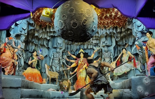A vibrant depiction of a mythological scene featuring various colorful statues and intricate details. Central figures include deities with multiple arms, a figure resembling an elephant, and other humanoid characters. The backdrop resembles a rocky cave with a large celestial object suspended above. Decorative elements and lighting add to the drama and complexity of the setting.