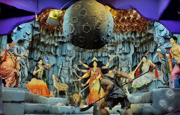 A vibrant depiction of a mythological scene featuring various colorful statues and intricate details. Central figures include deities with multiple arms, a figure resembling an elephant, and other humanoid characters. The backdrop resembles a rocky cave with a large celestial object suspended above. Decorative elements and lighting add to the drama and complexity of the setting.