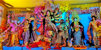 A vibrant and colorful display of Hindu deities in traditional attire. The central figure is a multi-armed goddess with a weapon, standing on a lion. Surrounding her are other deities, each with distinct postures and expressions, adorned with jewelry and floral decorations. The backdrop is richly decorated with flowers and fabric.