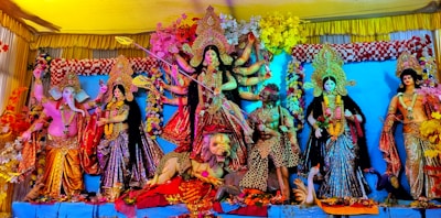 A vibrant and colorful display of Hindu deities in traditional attire. The central figure is a multi-armed goddess with a weapon, standing on a lion. Surrounding her are other deities, each with distinct postures and expressions, adorned with jewelry and floral decorations. The backdrop is richly decorated with flowers and fabric.