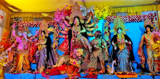 A vibrant and colorful display of Hindu deities in traditional attire. The central figure is a multi-armed goddess with a weapon, standing on a lion. Surrounding her are other deities, each with distinct postures and expressions, adorned with jewelry and floral decorations. The backdrop is richly decorated with flowers and fabric.
