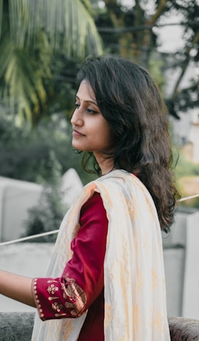 A young woman with long dark hair is wearing a maroon traditional outfit with detailed embroidery on the sleeve and a light shawl draped over her shoulder. She is outdoors, with a blurred background of greenery and structures, suggesting a tranquil setting.