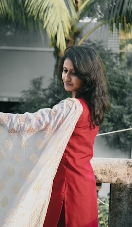 A woman with long, dark hair is wearing a red traditional outfit while holding a patterned shawl. She is standing on a balcony with greenery and a palm tree in the background, creating a natural setting.