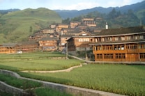 Traditional wooden houses are nestled on a lush, terraced mountainside, surrounded by greenery and overlooking vast rice paddies. The architecture features intricate wooden designs and sloped roofs, typical of rural Asian villages.