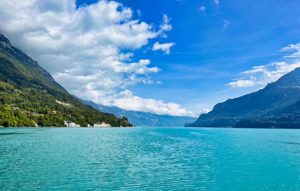 The image depicts a serene lake surrounded by mountains, with a clear blue sky above and fluffy white clouds below. The calm water reflects the surrounding landscape, creating a mirror-like effect that enhances the beauty of the scene. On one side of the lake, there is a small island covered in lush green vegetation, while on the other side, a larger island can be seen.

The mountains in the background are covered in trees and shrubs, adding depth to the landscape. The sky above is mostly clear with some clouds scattered across it, providing a beautiful backdrop for the lake and its surroundings.