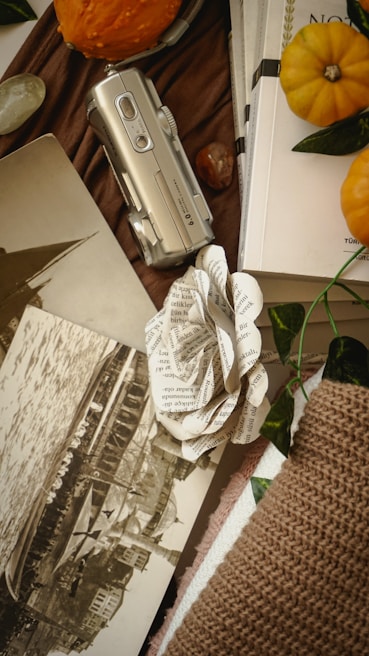 An arrangement of items includes a silver vintage camera, an old sepia-toned photograph depicting a waterfront scene, and a handcrafted paper flower made from printed text. There are pumpkins and books, suggesting an autumnal theme, and a textured brown fabric that adds a cozy feel.