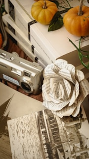 Several pumpkins, a vintage camera, a paper flower made from book pages, a stack of books, and an old photograph are arranged together. The image gives a warm and nostalgic tone with a touch of creativity through the handmade paper flower.
