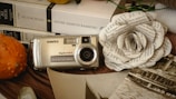 An antique digital camera is displayed on a table along with a few books and a paper flower made from printed text. The books by Victor Hugo can be seen clearly. To the left, there is a gourd with a spiky surface. The setup includes sepia-toned photographs giving a nostalgic feel. The items are arranged on a cloth.