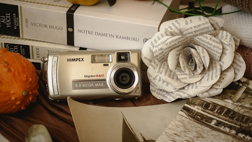 An antique digital camera is displayed on a table along with a few books and a paper flower made from printed text. The books by Victor Hugo can be seen clearly. To the left, there is a gourd with a spiky surface. The setup includes sepia-toned photographs giving a nostalgic feel. The items are arranged on a cloth.