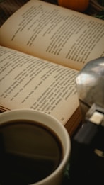 An open book with printed text creates a cozy reading atmosphere alongside a cup of coffee. A metallic object, possibly a coffee maker, is partially visible on the right.
