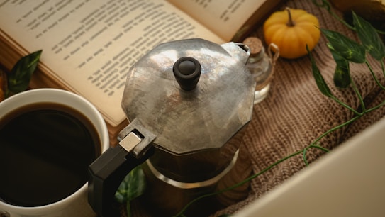 A vintage coffee maker sits on a textured surface beside a white cup filled with black coffee. An open book with visible text is placed in the background. Small decorative items, including a miniature pumpkin, a bottle, and green leaves, add a cozy and autumnal ambiance.