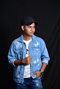 A young man wearing a distressed blue denim jacket with writing, paired with a white T-shirt and dark cap. He is looking off to the side with a thoughtful expression. He is also wearing a wristwatch and a black wristband. The background is a simple, dark curtain.