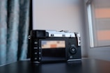 A digital camera with a retro design is resting on a dark surface near a window. The camera has a textured black and silver body with various buttons and a display screen on the back. The background features blue patterned curtains and part of a window frame, allowing natural light to illuminate the scene.