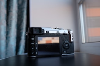 A digital camera with a retro design is resting on a dark surface near a window. The camera has a textured black and silver body with various buttons and a display screen on the back. The background features blue patterned curtains and part of a window frame, allowing natural light to illuminate the scene.