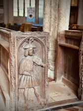 An intricately carved wooden panel features a medieval figure dressed in period attire, holding a shield. The panel is part of a pew in an old church, with stone columns and stained glass windows in the background.