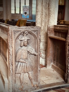 An intricately carved wooden panel features a medieval figure dressed in period attire, holding a shield. The panel is part of a pew in an old church, with stone columns and stained glass windows in the background.