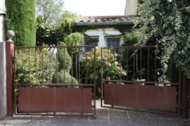 A rustic, metal gate partially open, revealing a lush garden area. The garden features a variety of green plants and bushes with some pink flowers. Behind the greenery, a small building with cream-colored walls and a tiled roof is visible. Ivy climbs over parts of the gate and house, enhancing the verdant setting. The scene suggests tranquility and natural beauty.