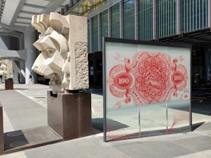 A modern architectural scene with geometric stone sculptures placed on dark pedestals, juxtaposed against a glass panel featuring a red decorative design reminiscent of banknote artwork. The area is bathed in sunlight, creating strong shadows.