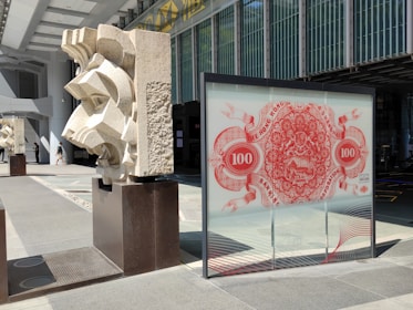 A modern architectural scene with geometric stone sculptures placed on dark pedestals, juxtaposed against a glass panel featuring a red decorative design reminiscent of banknote artwork. The area is bathed in sunlight, creating strong shadows.