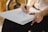 A person is sketching on a stack of papers using a pencil. Their hands are smudged with what appears to be charcoal or graphite. The setting includes a blurred background, suggesting an outdoor or well-lit area.