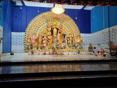 A vibrant and ornate display features a group of intricately decorated statues. The central figure is a goddess, flanked by other deities, set against a backdrop of colorful, symmetric patterns. The scene is adorned with floral arrangements and garlands, while offerings are placed on the floor.