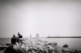 A black and white photograph capturing a group of people walking on a rocky pier extending into the sea. The scene includes distant lighthouses and a sign positioned along the pier. The photo conveys a sense of adventure and exploration.