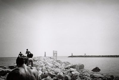 A black and white photograph capturing a group of people walking on a rocky pier extending into the sea. The scene includes distant lighthouses and a sign positioned along the pier. The photo conveys a sense of adventure and exploration.