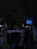 Inside of a dimly lit train compartment with sleeping berths, luggage stored on the upper racks, and a blue digital display showing seat numbers. The atmosphere seems cramped and utilitarian.