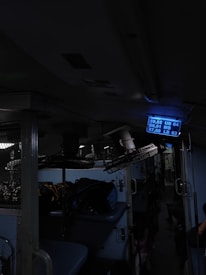 Inside of a dimly lit train compartment with sleeping berths, luggage stored on the upper racks, and a blue digital display showing seat numbers. The atmosphere seems cramped and utilitarian.