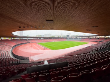 A large, open-air stadium with a modern design, showcasing a well-maintained green sports field surrounded by a red running track. The seating areas are empty, with rows of red seats extending around the field. The curvature of the wooden ceiling adds architectural interest, and the view includes surrounding buildings and a pale sky.