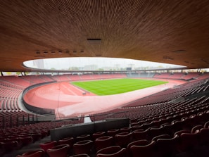 A large, open-air stadium with a modern design, showcasing a well-maintained green sports field surrounded by a red running track. The seating areas are empty, with rows of red seats extending around the field. The curvature of the wooden ceiling adds architectural interest, and the view includes surrounding buildings and a pale sky.