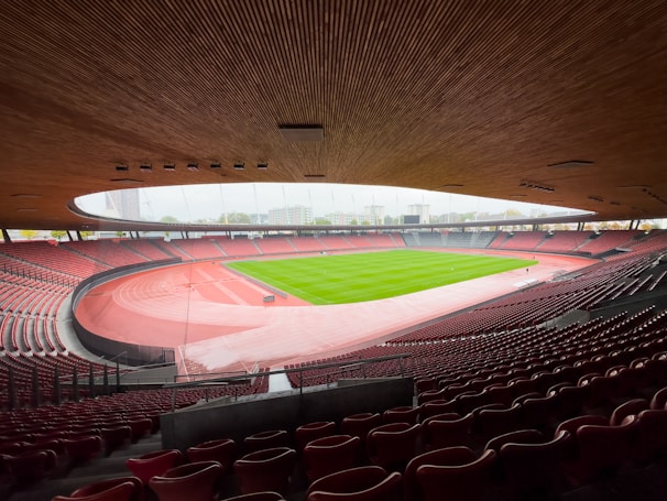 A large, open-air stadium with a modern design, showcasing a well-maintained green sports field surrounded by a red running track. The seating areas are empty, with rows of red seats extending around the field. The curvature of the wooden ceiling adds architectural interest, and the view includes surrounding buildings and a pale sky.