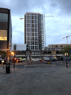 A tall modern building under construction dominates the scene, with cranes visible against a cloudy sky. Surrounding the central structure are various smaller buildings and parked vehicles. Streetlights illuminate the foreground, and there is a gated area leading to a parking lot.