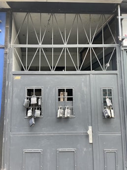 A gray metal gate with multiple lockboxes hanging from small barred windows. The upper section of the gate has a series of triangular metal beams. The presence of these lockboxes suggests security and controlled access.