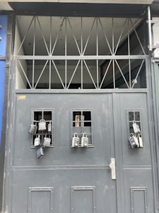A gray metal gate with multiple lockboxes hanging from small barred windows. The upper section of the gate has a series of triangular metal beams. The presence of these lockboxes suggests security and controlled access.