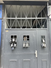 A gray metal gate with multiple lockboxes hanging from small barred windows. The upper section of the gate has a series of triangular metal beams. The presence of these lockboxes suggests security and controlled access.
