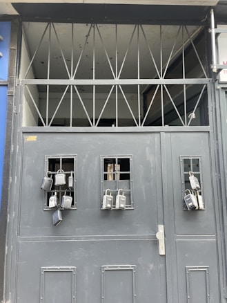 A gray metal gate with multiple lockboxes hanging from small barred windows. The upper section of the gate has a series of triangular metal beams. The presence of these lockboxes suggests security and controlled access.