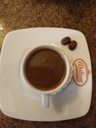 A cup of coffee on a white saucer with the brand name 'Delice' inscribed on it is placed on a stone-textured surface. Two chocolate pieces resembling coffee beans are positioned beside the cup.