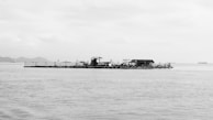 A solitary floating structure positioned in a vast body of water, possibly an aquaculture platform or fisherman's hut. The horizon features distant landmasses and ships, under a cloudy sky.